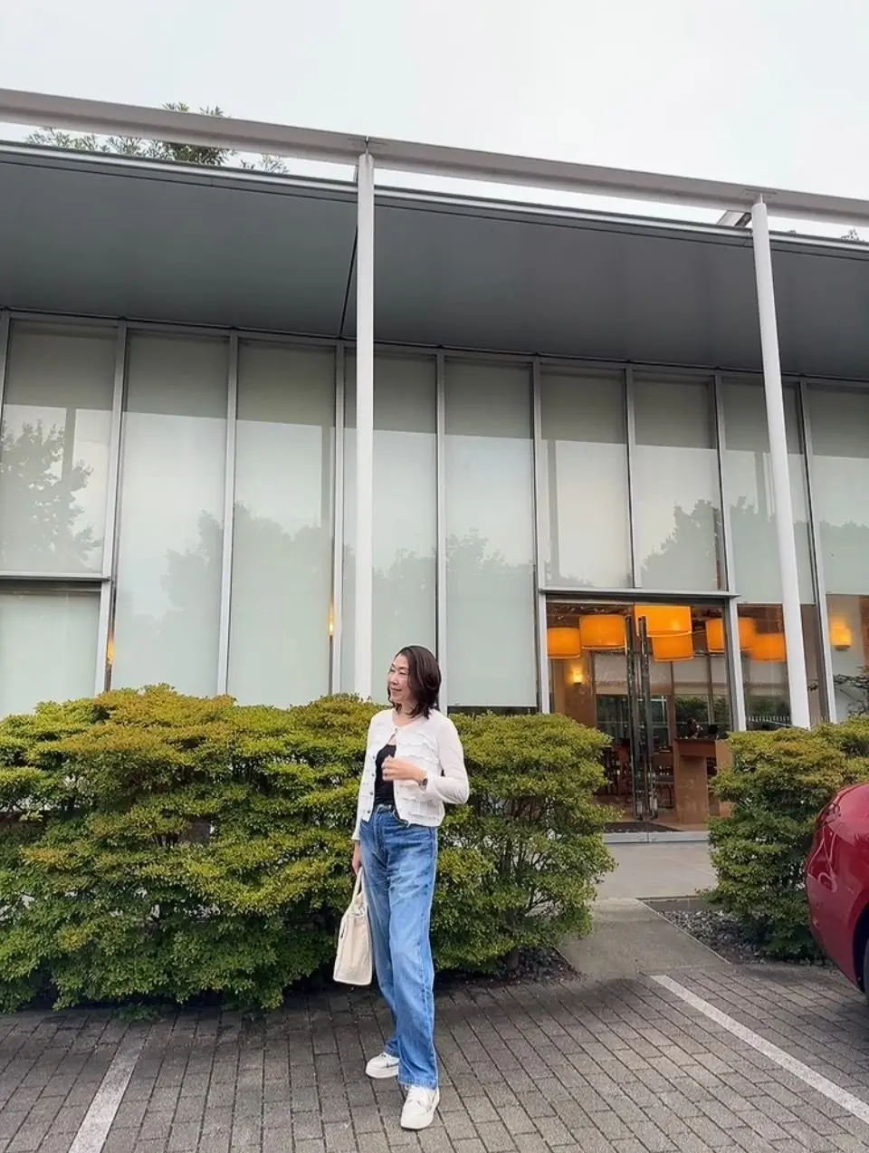 白いジャケットにデニムの女性が、グレーのタイル敷きの地面に立っている。背景には、白い壁と大きな窓のある近代的な建物。手には飲み物らしきもの、肩にはバッグをかけている。