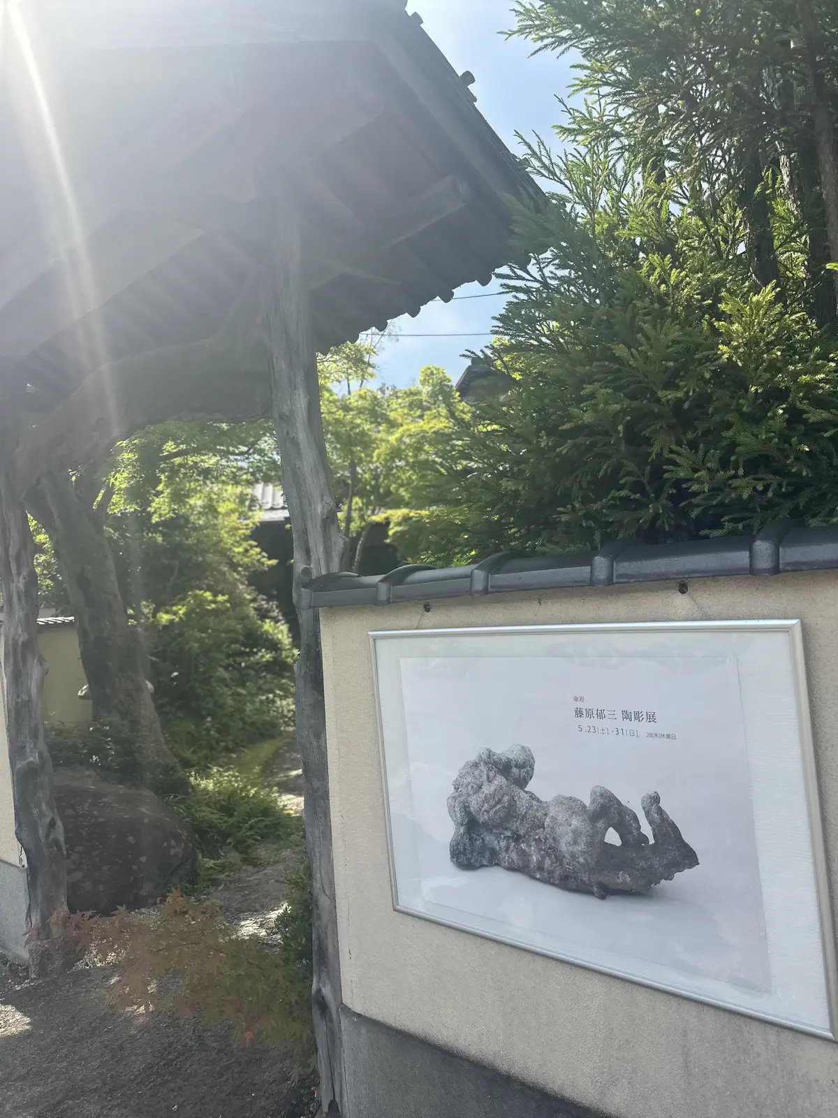 屋外の壁に展示された石像のポスター、木造建築と緑豊かな庭園が背景にある