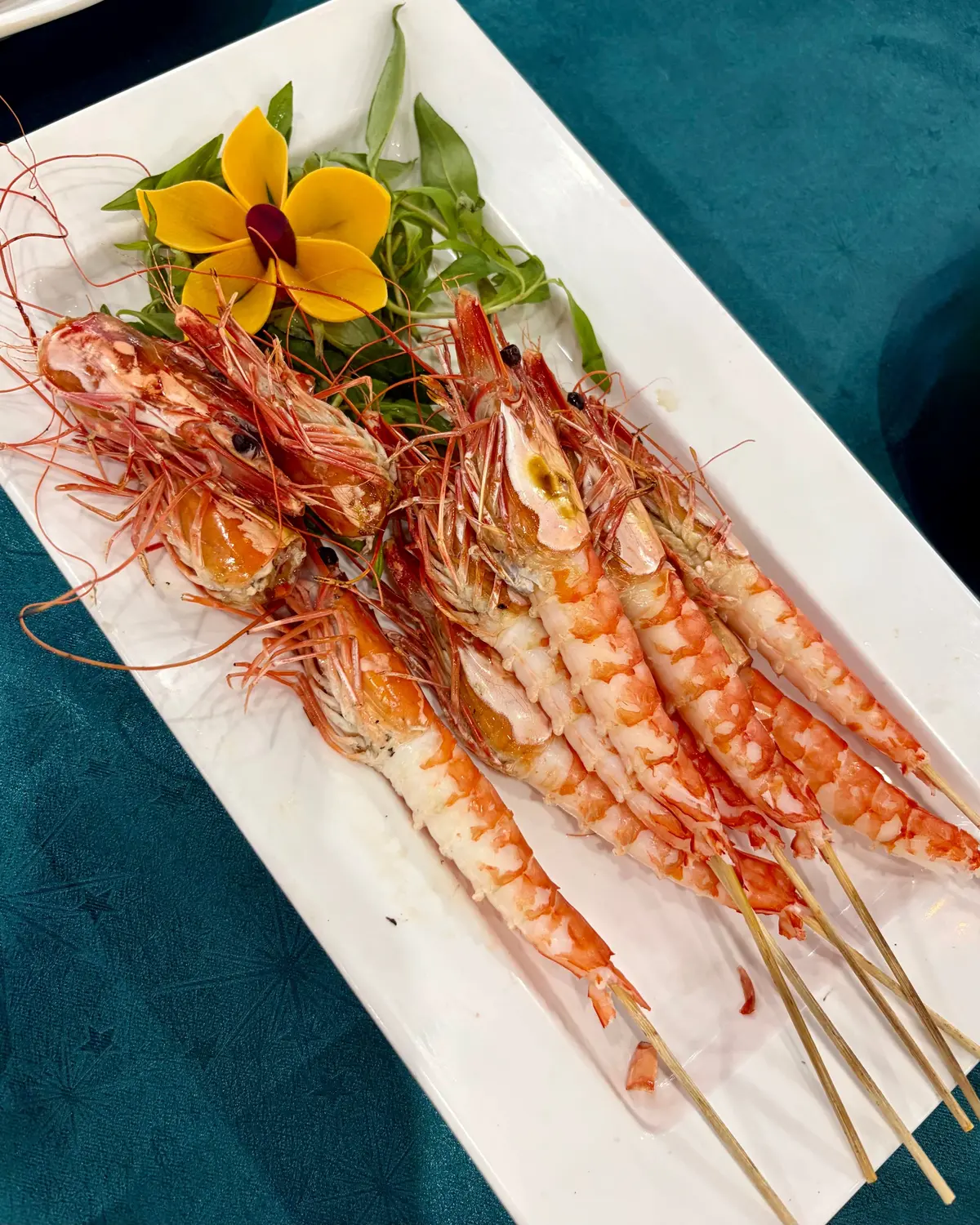 白い長方形のプレートに、串に刺したエビが複数並び、緑の葉野菜と黄色い花が添えられている。