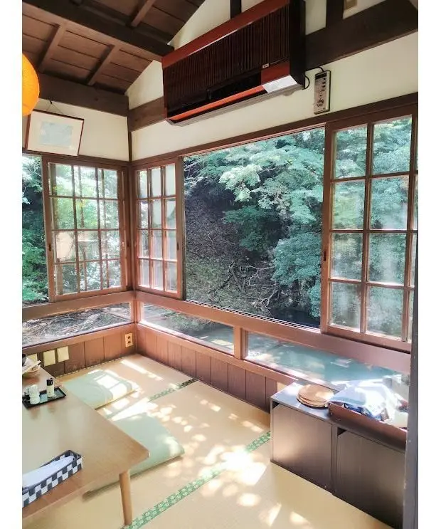 木造の部屋で、窓の外には緑豊かな木々が生い茂る渓流が見える。畳敷きの床に座椅子と机が置かれ、窓辺には木製のベンチがある。天井にはエアコンが設置されている。