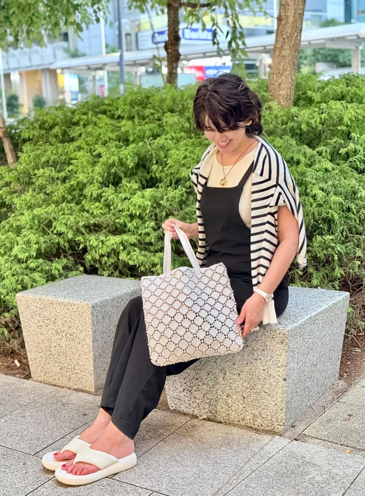 黒いパンツとサスペンダー付きの黒いトップスに、白黒ストライプの羽織りものを着た女性が、白いサンダルで座っている。手には白いトートバッグを持ち、背景には緑の植栽と都市の建物が見える。