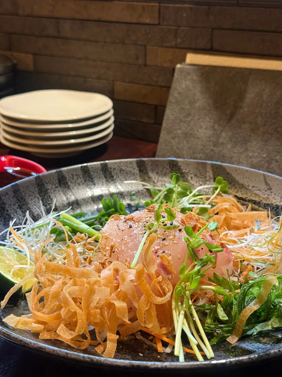 グレーの器に盛り付けられた、赤身のマグロと野菜の料理。カリカリのフライドオニオン、マイクログリーン、千切り野菜が彩りを添える。背景には皿の山とメニューがある。