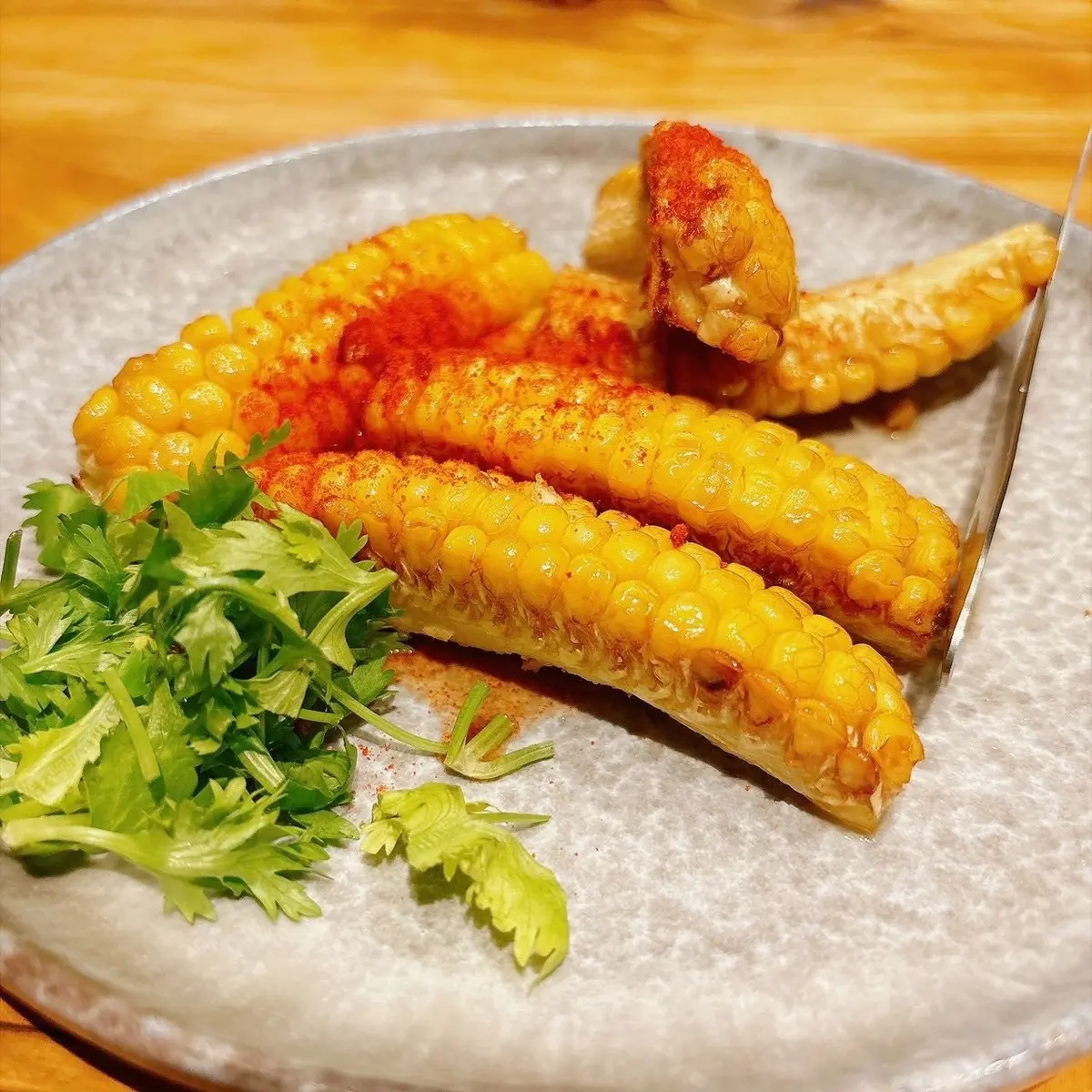 グレーの皿に盛り付けられた、赤パプリカを振ったコーン4本。葉の多い緑のハーブが添えられ、ナイフが置かれている。
