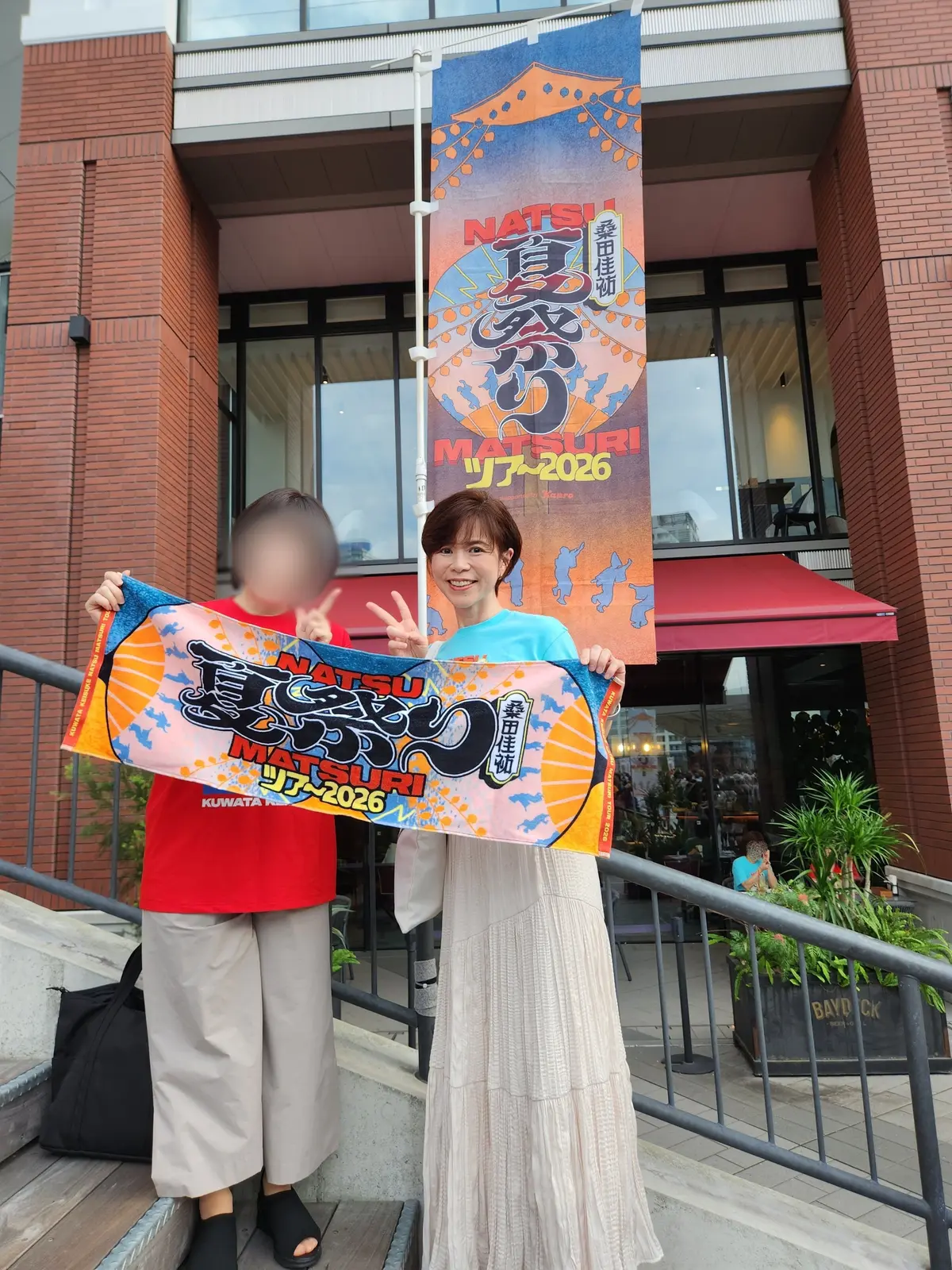 建物の前で、女性と少年が夏の祭り（Natsu Matsuri）の旗を持ち笑顔を見せている。旗にはオレンジと青のグラデーションで装飾された文字が描かれている。
