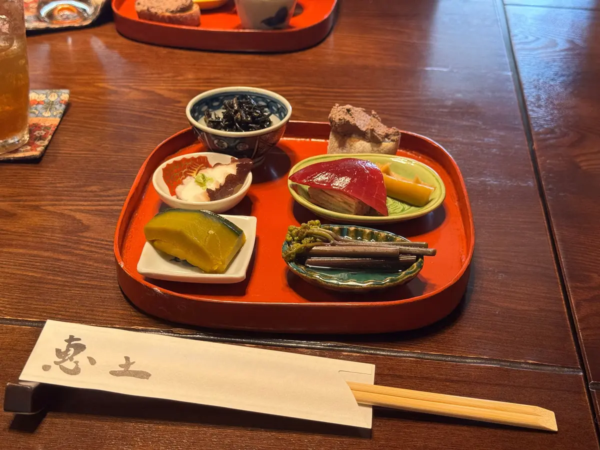 オレンジのトレイに、ワサビ入りの刺身、赤いカボチャの煮物、黄色の野菜の盛り合わせ、海藻の入った小鉢、ワカメと枝豆の小皿、焼き魚の切り身が並んでいる
