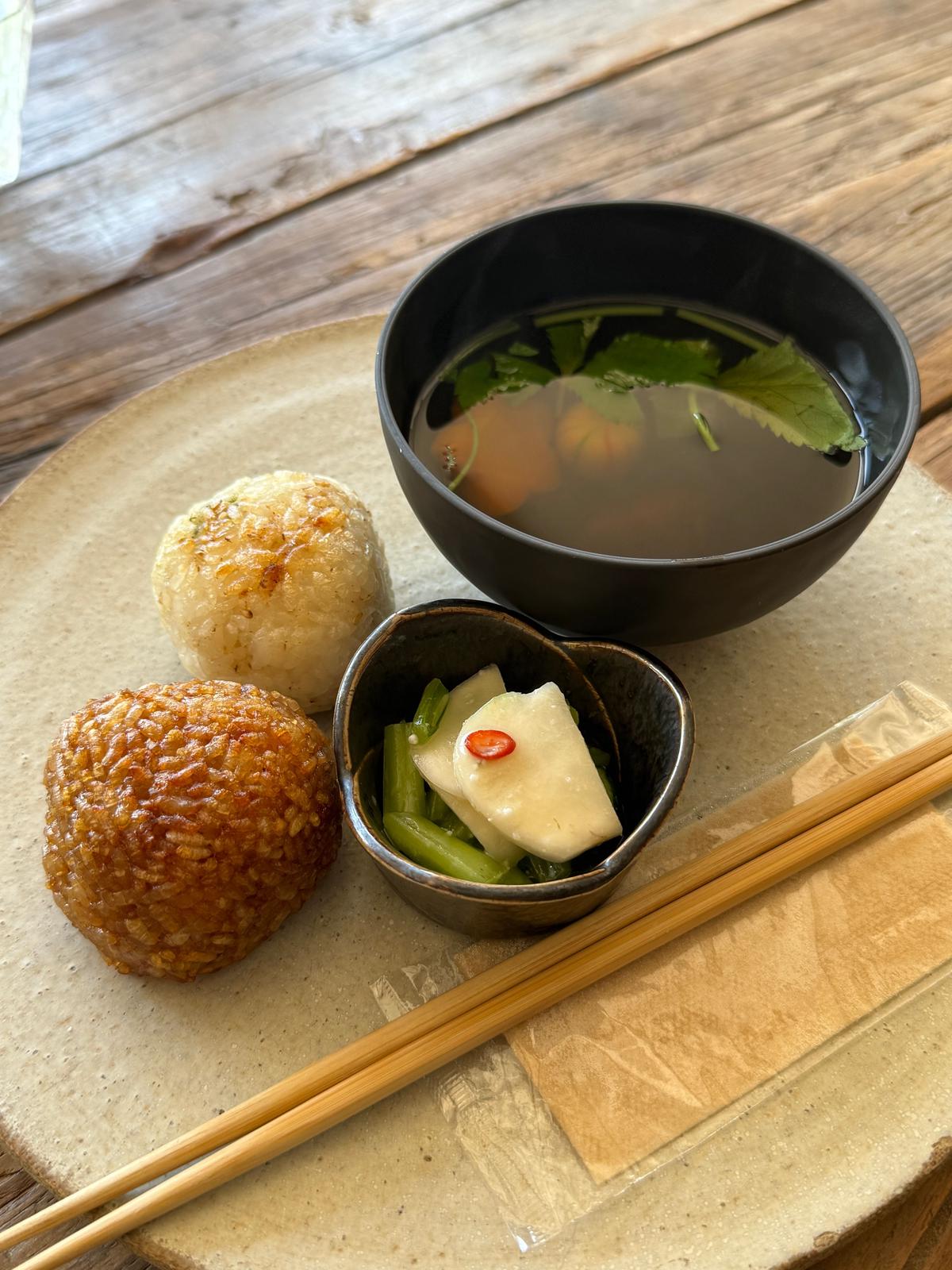 木製のテーブルに置かれた、和食の盛り合わせ。右には黒い汁椀に入ったスープ、左下には茶色いおこわ、左上には白くて丸いお餅。手前には小鉢に入った、緑色の野菜と白い切り身、赤い唐辛子。竹製の箸が添えられている。