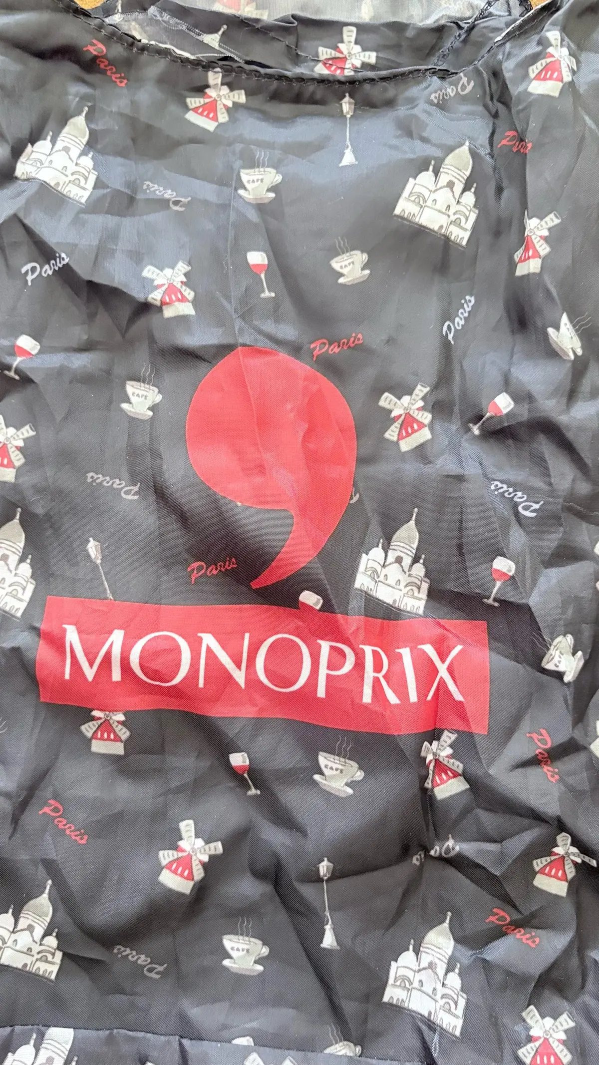 黒い背景にエッフェル塔やカフェのイラストが散りばめられた生地。中央に赤い丸と「MONOPRIX」のロゴが白文字で配置されている。