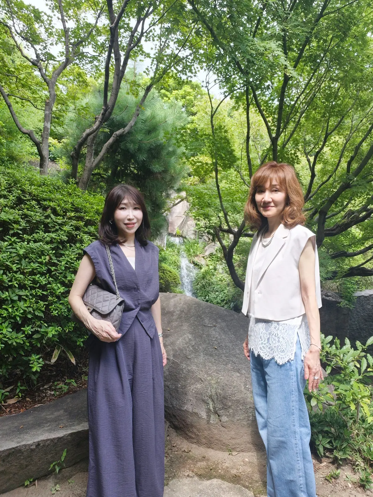 緑豊かな庭園を背景に、2人の女性が立っている写真。左は紫色のワンピース、右は白いジャケットとレースのトップスにデニムを着用。それぞれが笑顔でこちらを見ている。