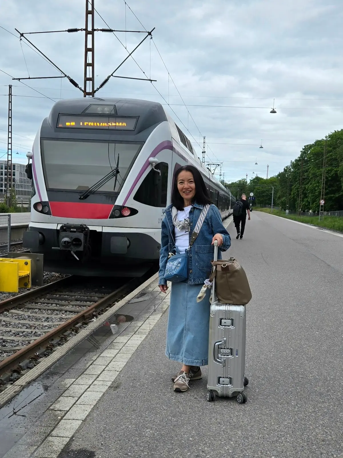 駅のホームに立つ女性。白い髪にデニムのジャケット、スカート、スニーカーを着用。シルバーのキャリーケースとバッグを持ち、笑顔でこちらを見ている。背景にはピンクと紫のラインが入った電車。