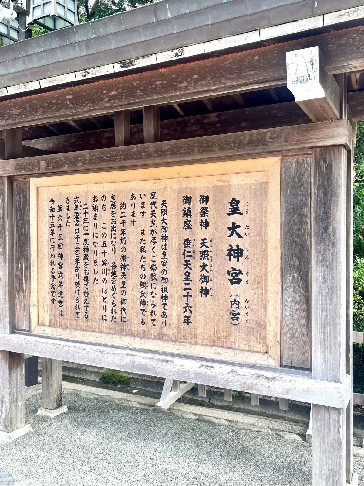 木造の鳥居の内部に設置された、木製の板に書かれた日本語の案内板。右上に小さな額縁に入った絵が飾られている。案内板には「皇大神宮（内宮）」の文字と、神楽神、天照大神、御鎮座垂仁天皇六年などの文字が記載されている。