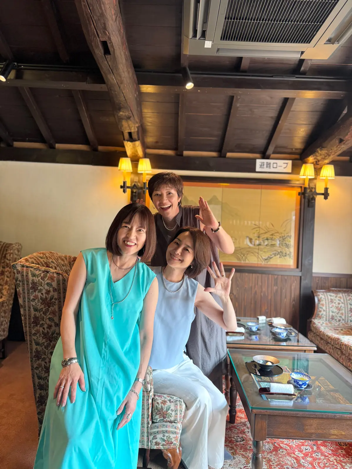 木造梁の天井がある部屋で、3人の女性が笑顔で写っている。左から青いワンピースを着た女性、白いシャツを着た女性、グレーの服を着た女性。中央の女性は手を挙げており、背景には水墨画が飾られている。