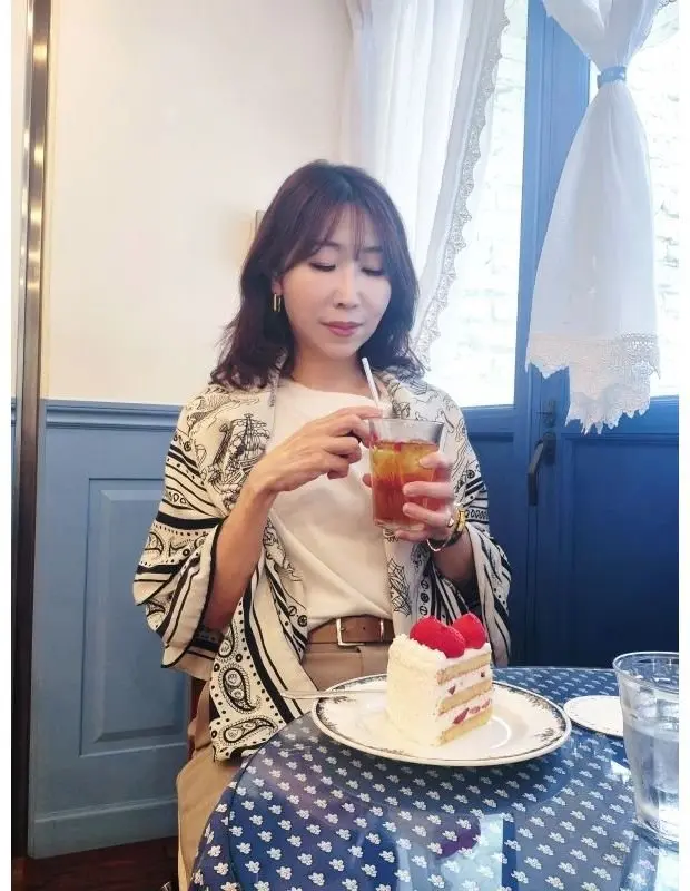女性が白いテーブルクロスに敷かれたテーブルで、ストローを使って飲み物を飲みながら、ラズベリーをトッピングした白いケーキを前にしています。