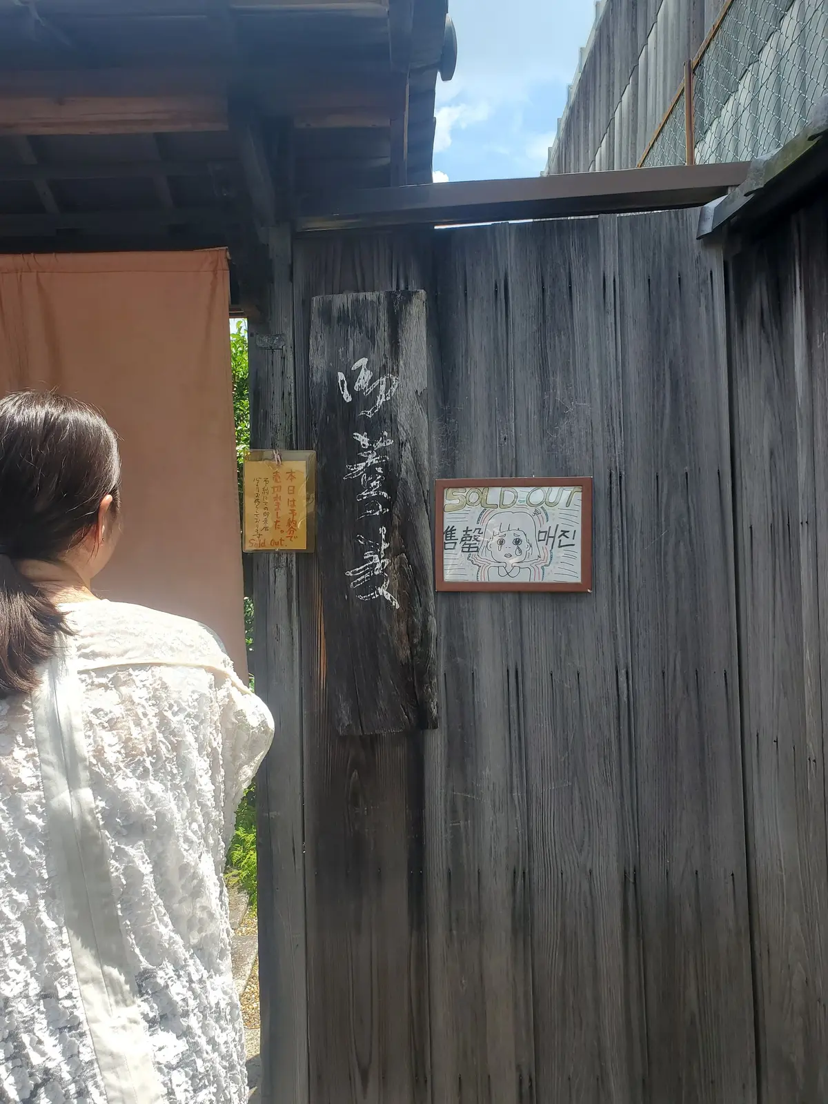 木製の小屋の扉に「SOLD OUT」と書かれた看板と、猫のイラストが描かれている。扉の横にはオレンジ色のカーテンがかかり、女性がこちらを向いている。