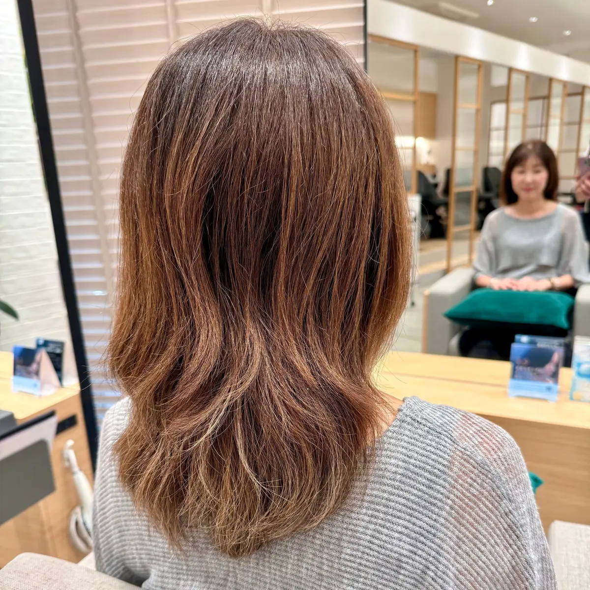肩下丈のゆるやかなウェーブヘア。根元は濃いブラウン、毛先にかけて明るいオレンジブラウンに変化。光の当たり方でゴールドの輝きが見える。後ろ姿で、灰色のニットを着用。