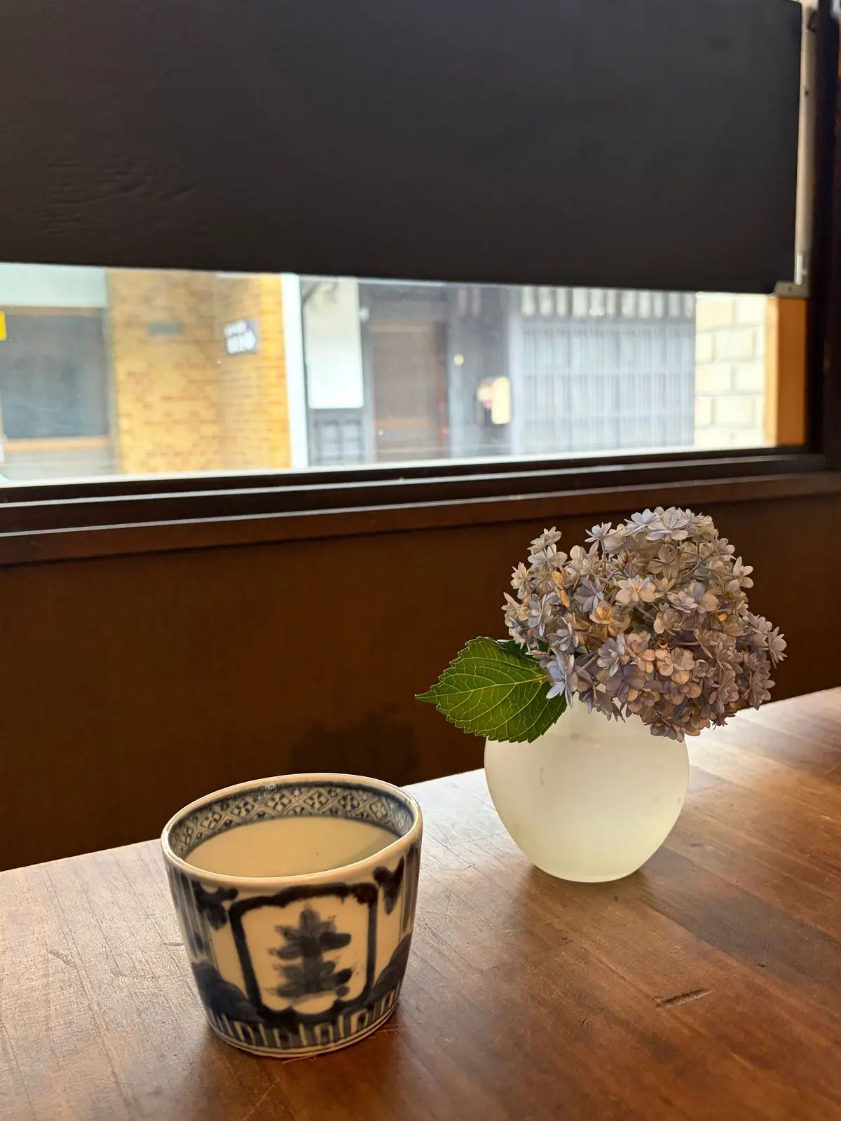 木のテーブルに青と白の模様のコーヒーカップと、紫の花が咲く花瓶が置かれている。窓の外に建物が見える。