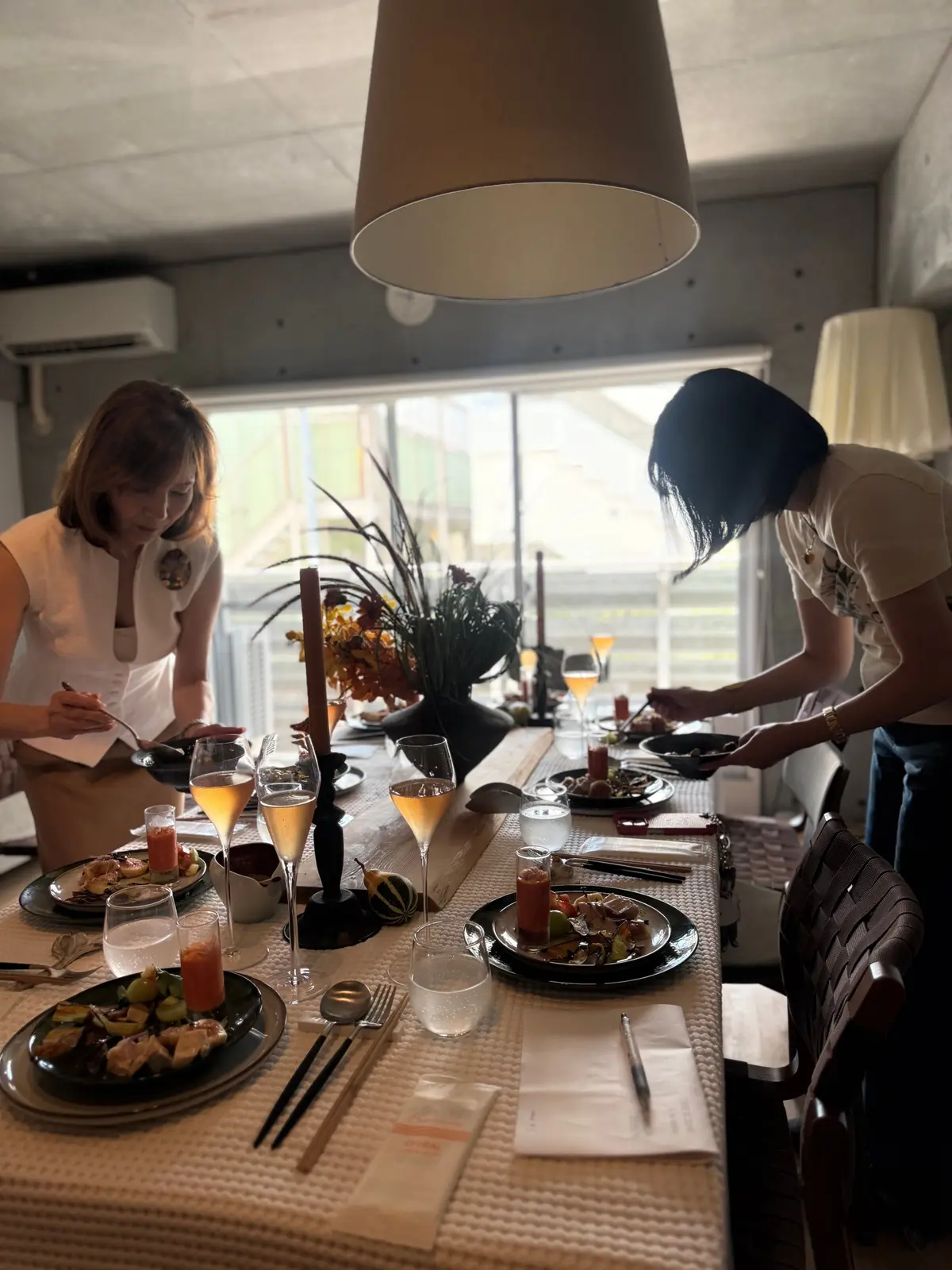 長テーブルを挟んで、女性2人が食事を楽しんでいる様子。テーブルには盛り付けられた料理とグラスが並び、中央には花瓶が置かれている。照明器具が吊り下げられ、窓から自然光が差し込んでいる。