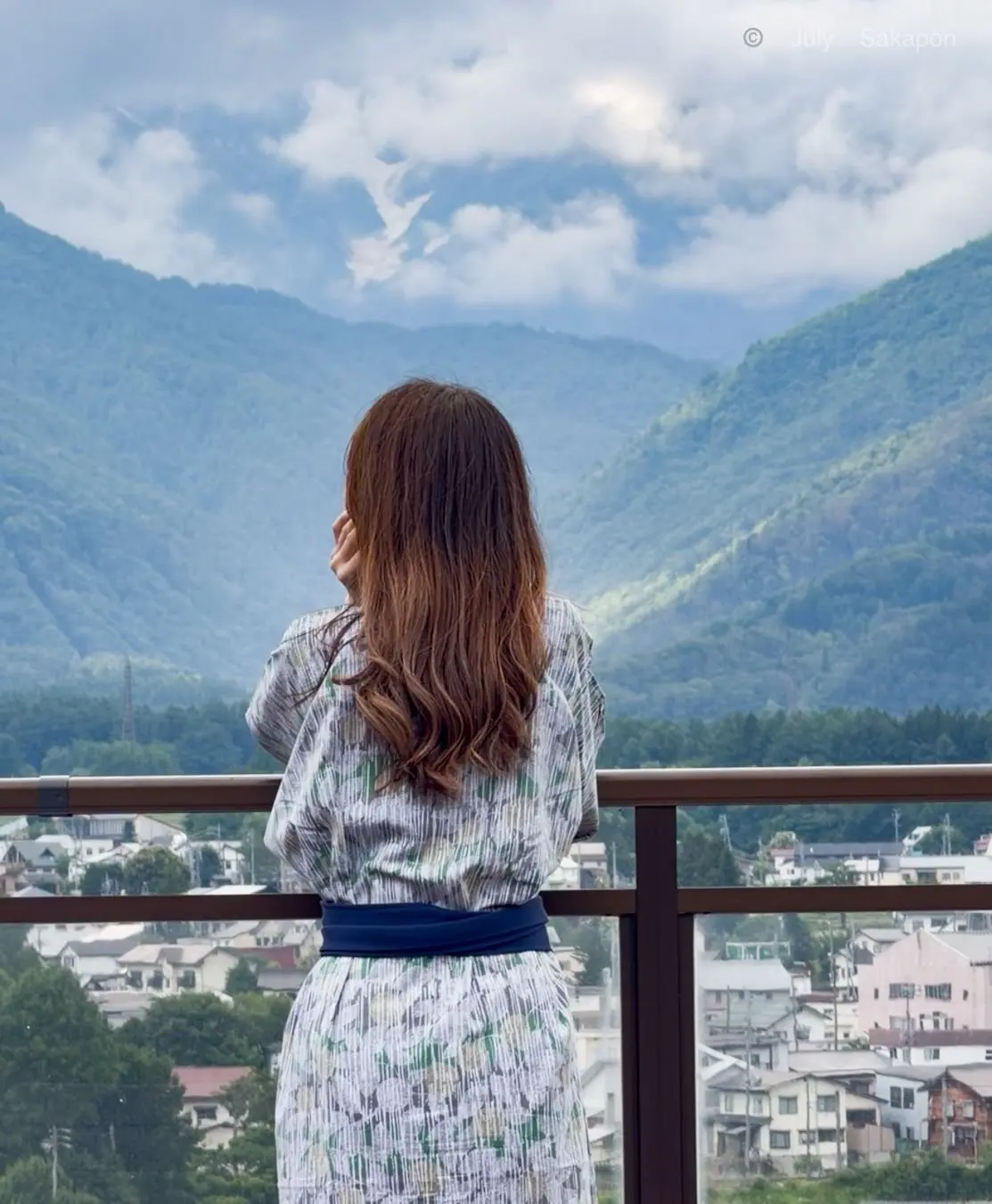 女性が手で耳を覆い、バルコニーの手すり越しに山並みと住宅地を眺めている