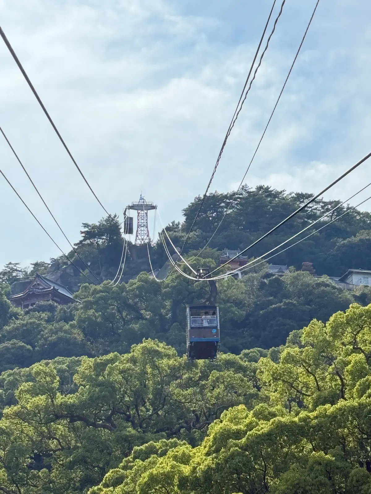 緑豊かな丘陵地帯をロープウェイが横断。頂上には白い塔のような建造物が見える。青空に電線が伸びている。広島、尾道で、尾道観光。尾道三部作の舞台巡り。