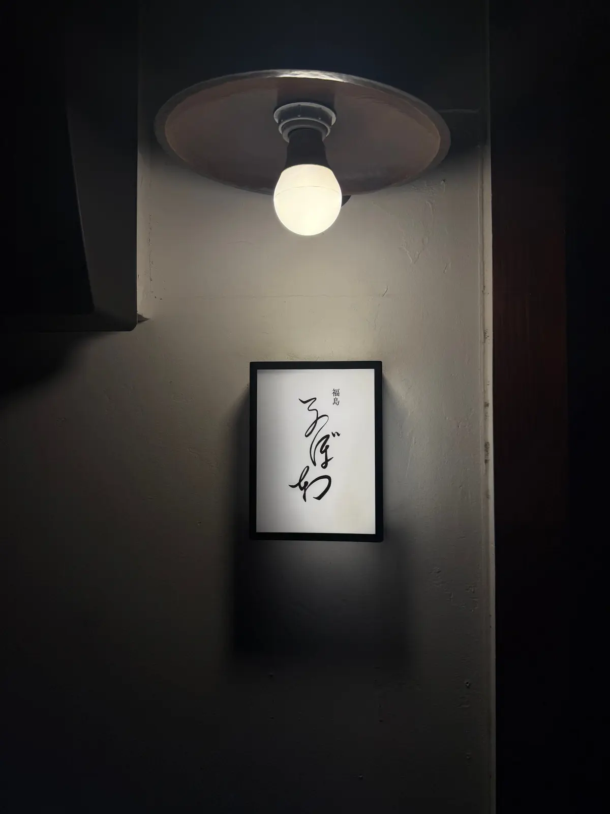 壁に掛けられた黒縁の絵画。白地に黒の線で描かれた抽象的な模様が描かれている。上部に照明器具があり、光が絵画を照らしている。背景は暗く、壁の質感が見える。