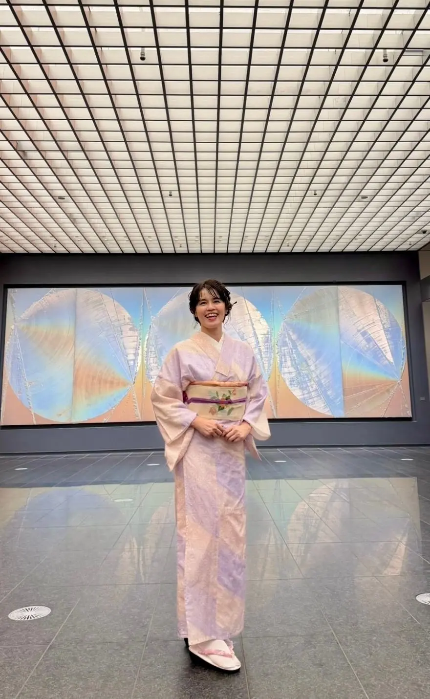 淡い紫色の着物を着た女性が、美術館のような空間で微笑んでいる。背景には抽象的な模様の絵画、天井は格子状の照明が並んでいる。