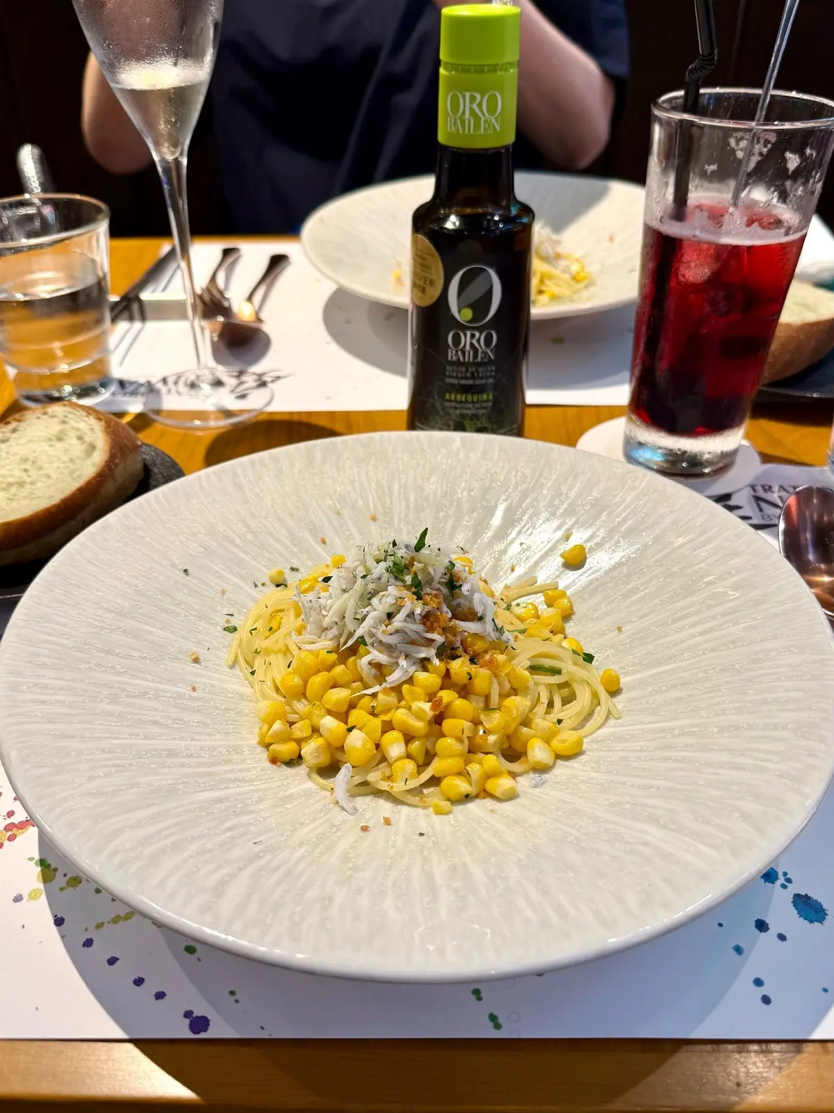 表参道レストラン、イタリアンレストラン庭、白いプレートに、トウモロコシとスパゲッティが盛り付けられ、チーズとハーブがトッピングされている。背景にはワイングラス、オリーブオイルのボトル、飲み物とスプーンが置かれている。