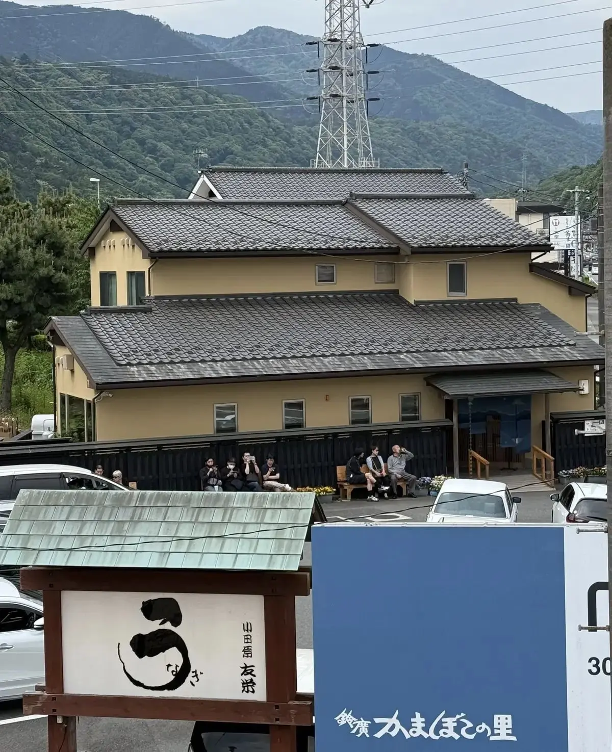 山間の風景の中にある、ベージュ色の2階建ての伝統的な日本家屋。軒先に人々が座り、建物前には車両が停車している。手前に青い看板があり、黒い円のマークと日本語の文字が書かれている。