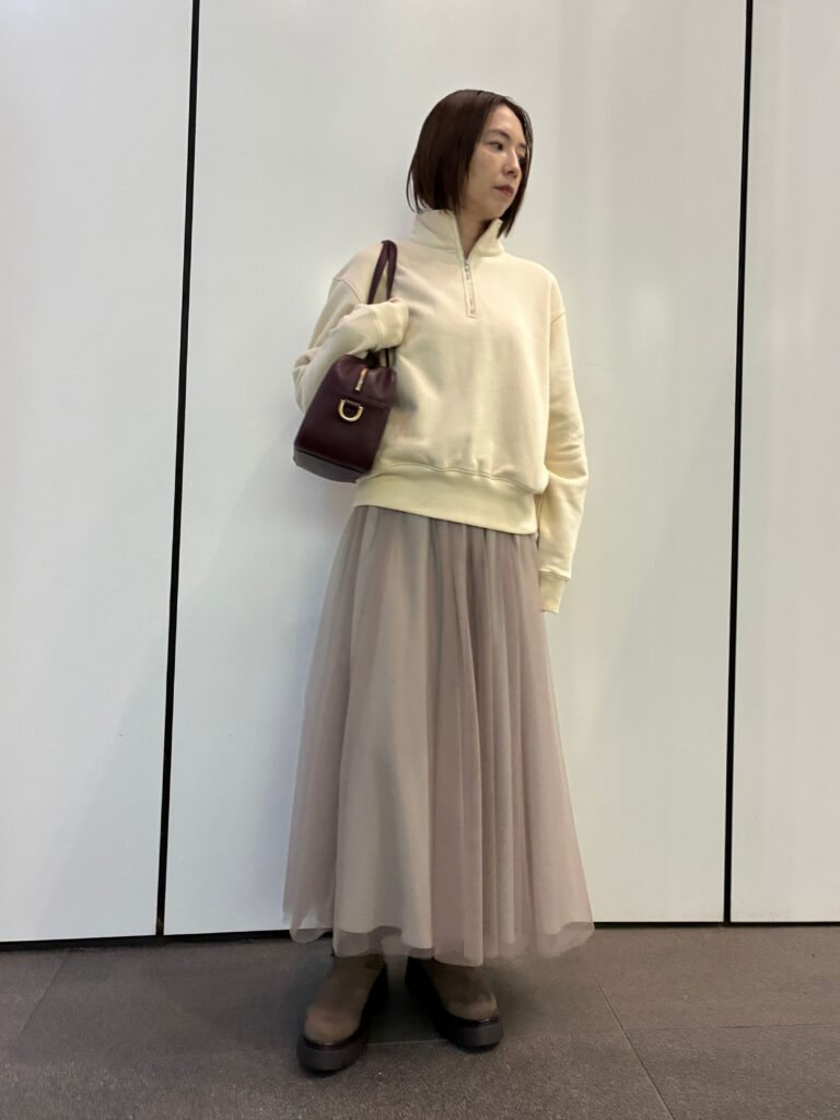 白のジップアップスウェットに、ベージュのチュールスカート、茶色のローヒールパンプスを合わせた女性。茶色のハンドバッグを肩にかけ、白い壁の前で立っている。