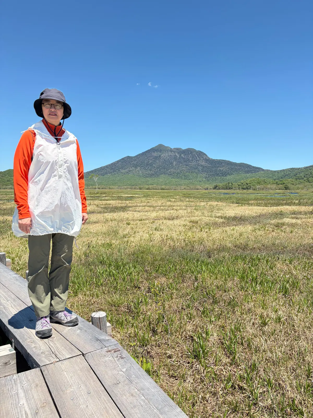 女性が木製の歩道に立ち、広大な湿地と山を背景にしています。女性はグレーの帽子、オレンジの長袖、緑のズボン、白いベストを着ています。晴れた空と緑豊かな山が遠くに見えます。