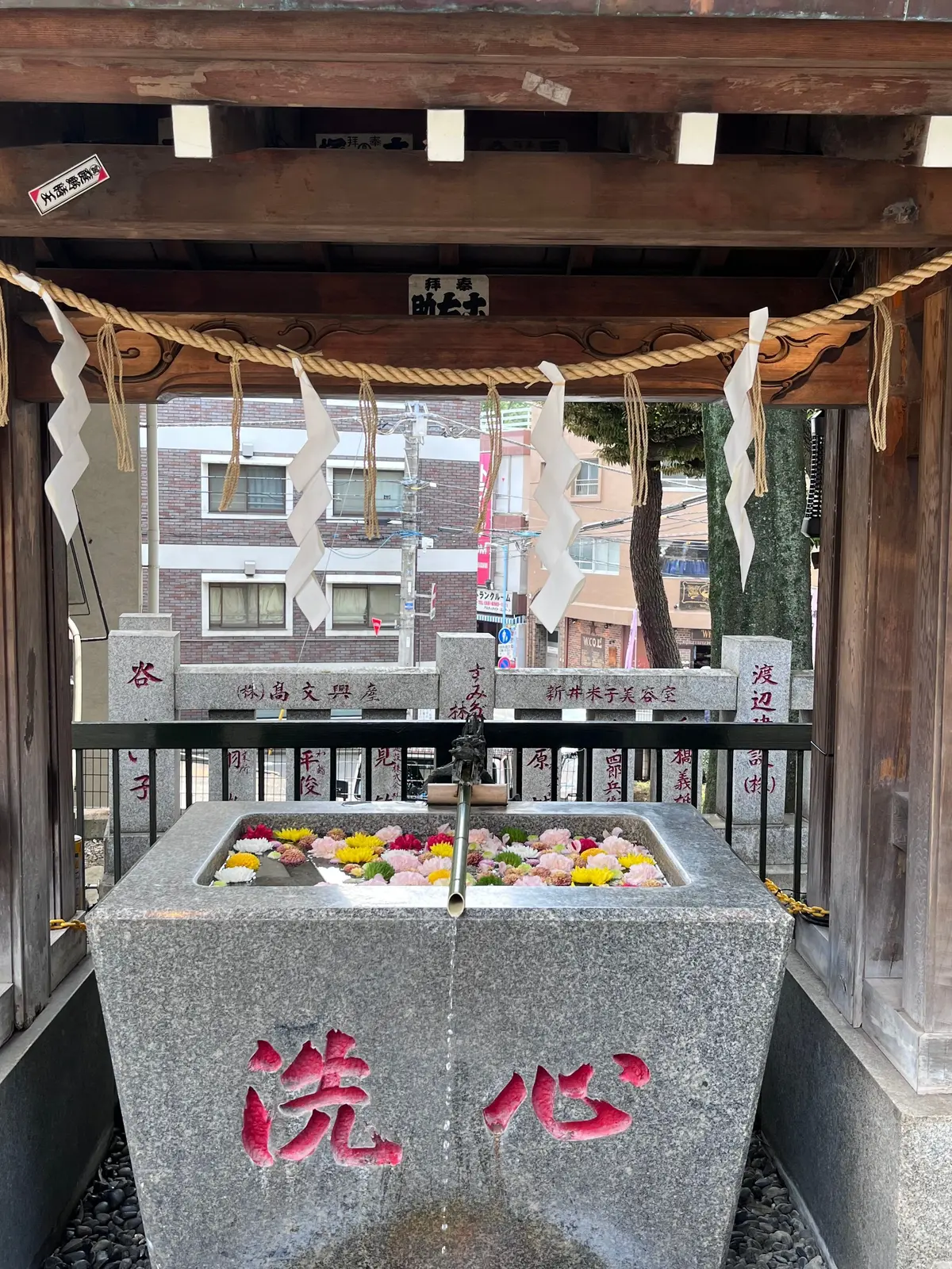 石造りの手水舎。中央に水が流れ出る蛇口があり、花が飾られている。屋根には提灯と白い紙垂が吊るされ、背景に街並みが見える。
