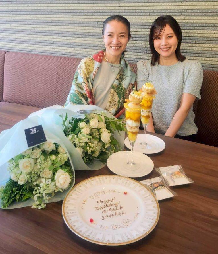 二人の女性が笑顔で並び、花束とケーキが置かれたテーブルに座っている。