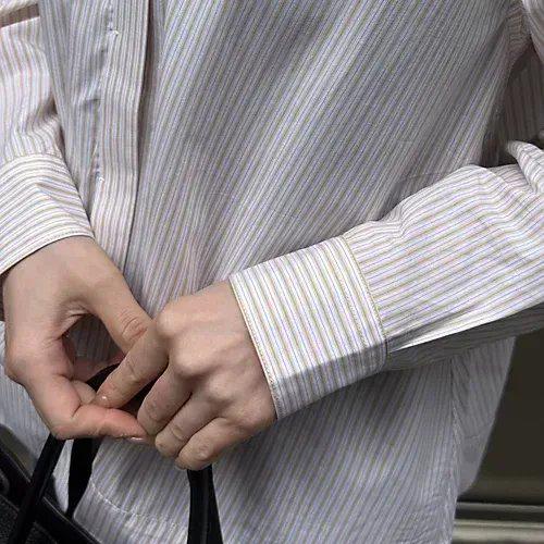 ベージュと白の細いストライプ柄のシャツを着た腕。ボタンがいくつか開いており、黒いバッグを持っている。