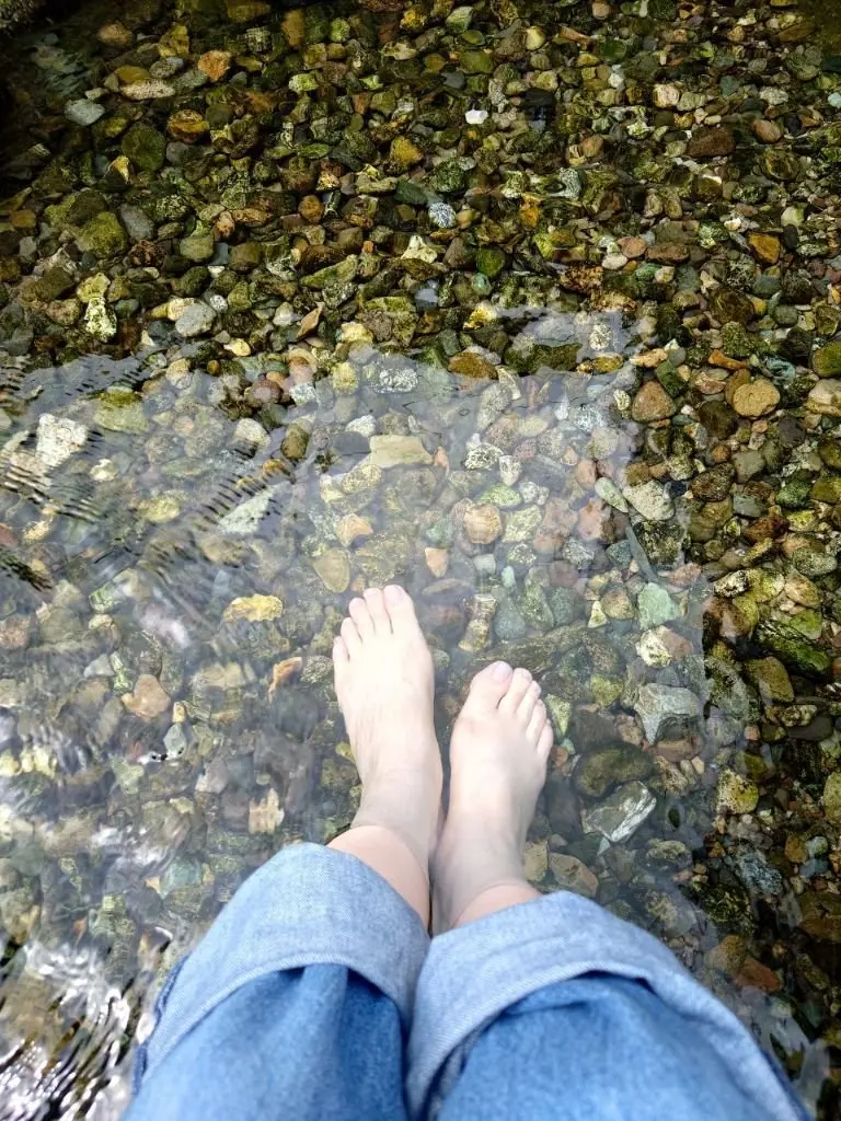 水辺に置かれた足。透明度の高い水底には、大小さまざまな石が敷き詰められている。足は水に浸かり、水面がわずかに波紋になっている。青いジーンズを履いている。