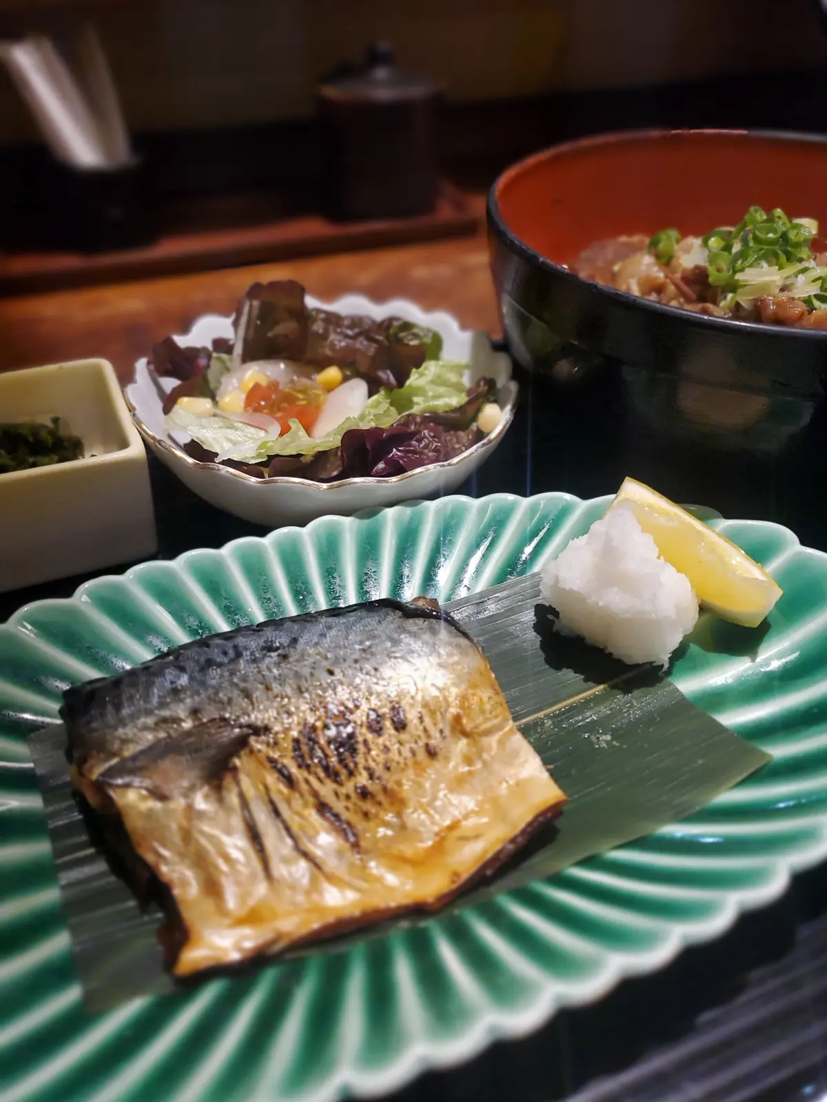 緑色の波紋模様の皿に、焼きたてのサンマが乗っている。隣にはレモンと塩の山。背景にはサラダとご飯もの。