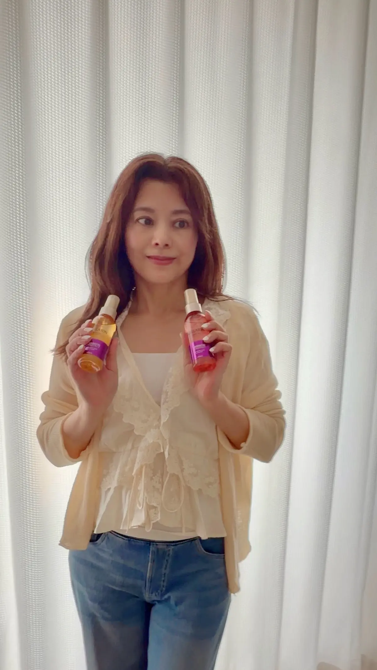 女性が、黄色いカーディガンを着用し、両手にウエラのヘアオイルのボトルを持つ。背景は白いカーテン。