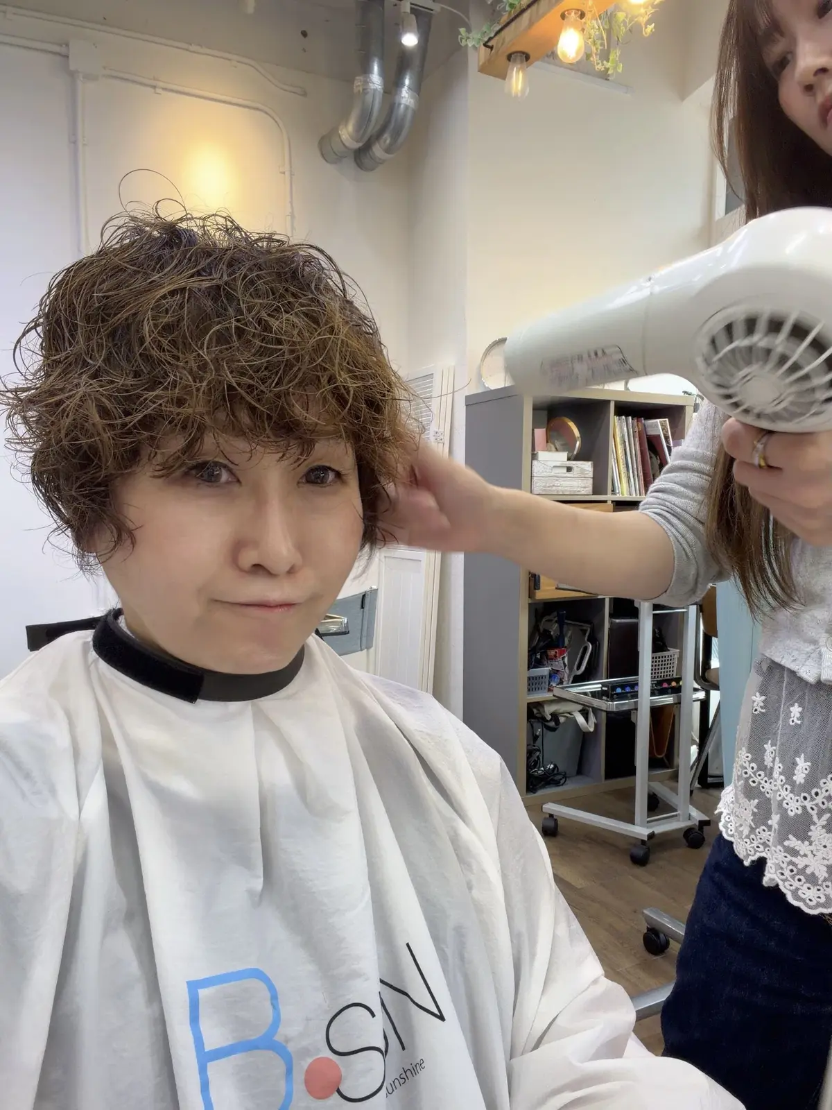 美容室でカットを受けた若い男女。肩までの長さの暗いカールヘアで、白いカットクロスを着用。背景には丸い鏡と、BSNのロゴが入った装飾がある。