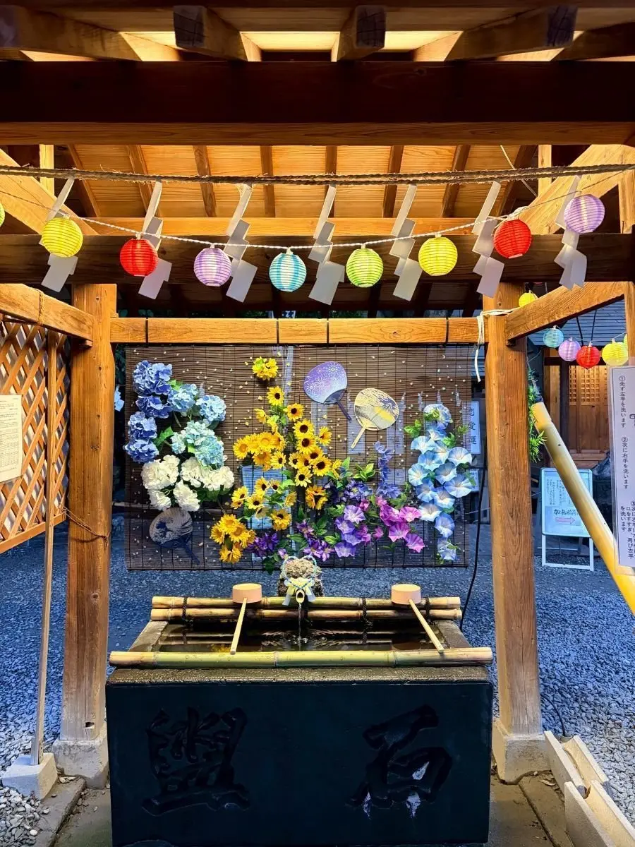 木造の簡素な祠に、花と扇子を飾った手水舎。竹製の台に水が流れ、両側に木製の柄が設置されている。屋根には多色の提灯が吊るされ、背景には竹製の格子と看板が見える。