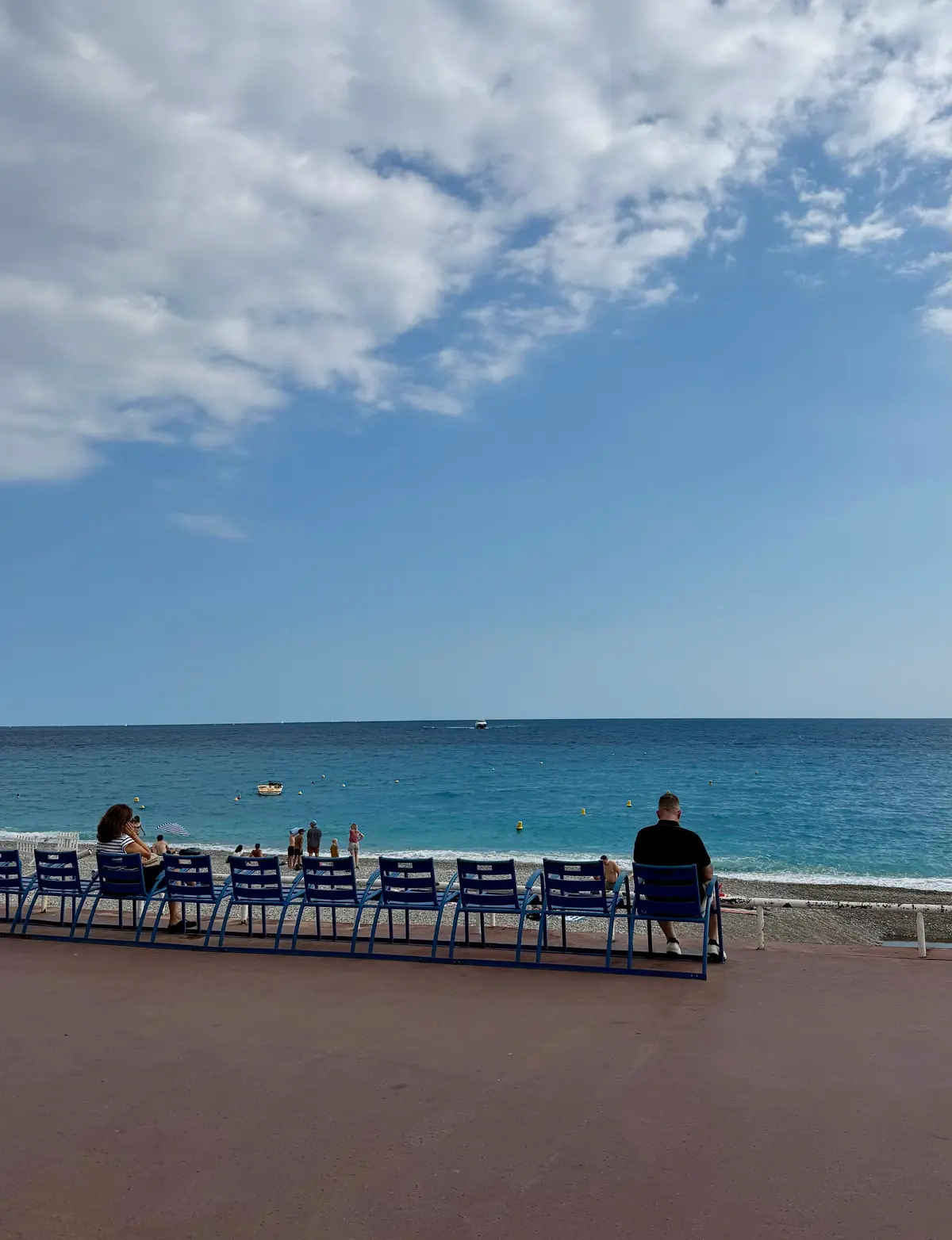 青い椅子が並ぶビーチ。穏やかな青い海と、白と青の雲空が広がる。手前には赤茶色の歩道が続く。