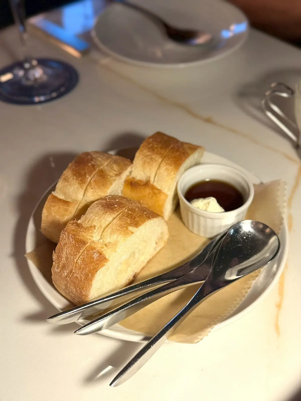 白いプレートに切られたパンと、小さなボウルにディップソースとバターが置かれています。銀のフォークとナイフがパンにかかっています。