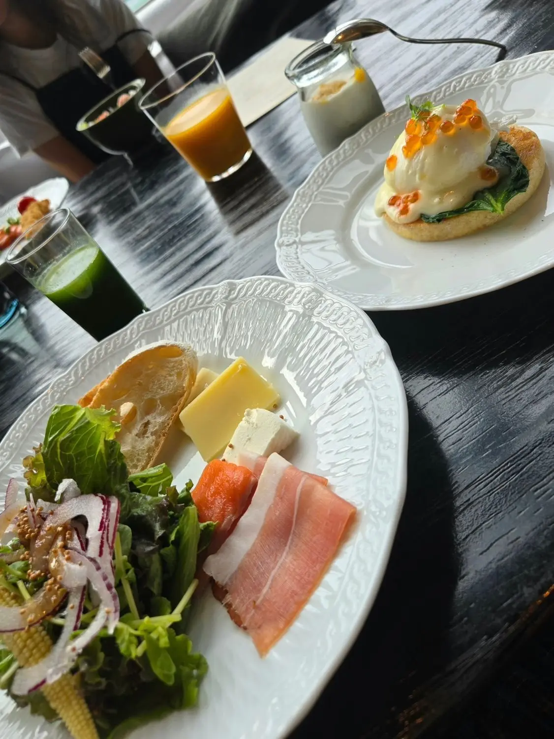 黒い木製のテーブルに置かれた、様々な料理と飲み物の写真。左には緑色のジュース、右にはオレンジ色のジュースと白い器に入った飲み物、奥にはコーヒーが見える。