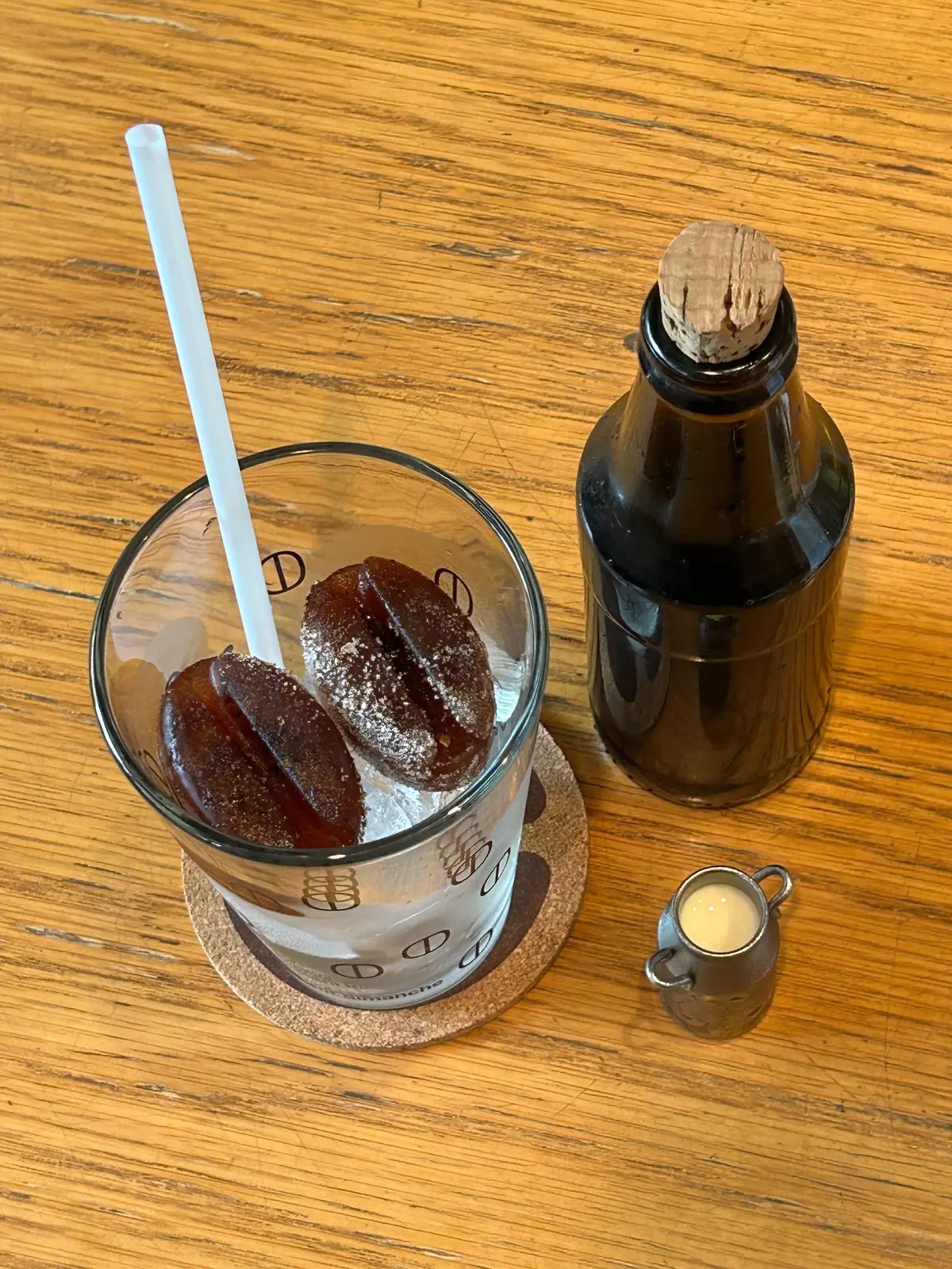 木製のテーブルに置かれた、透明なグラスに入ったコーヒー風味のグミと、それに合う飲み物。右には黒い瓶、下にはミルクが入った小さな容器。