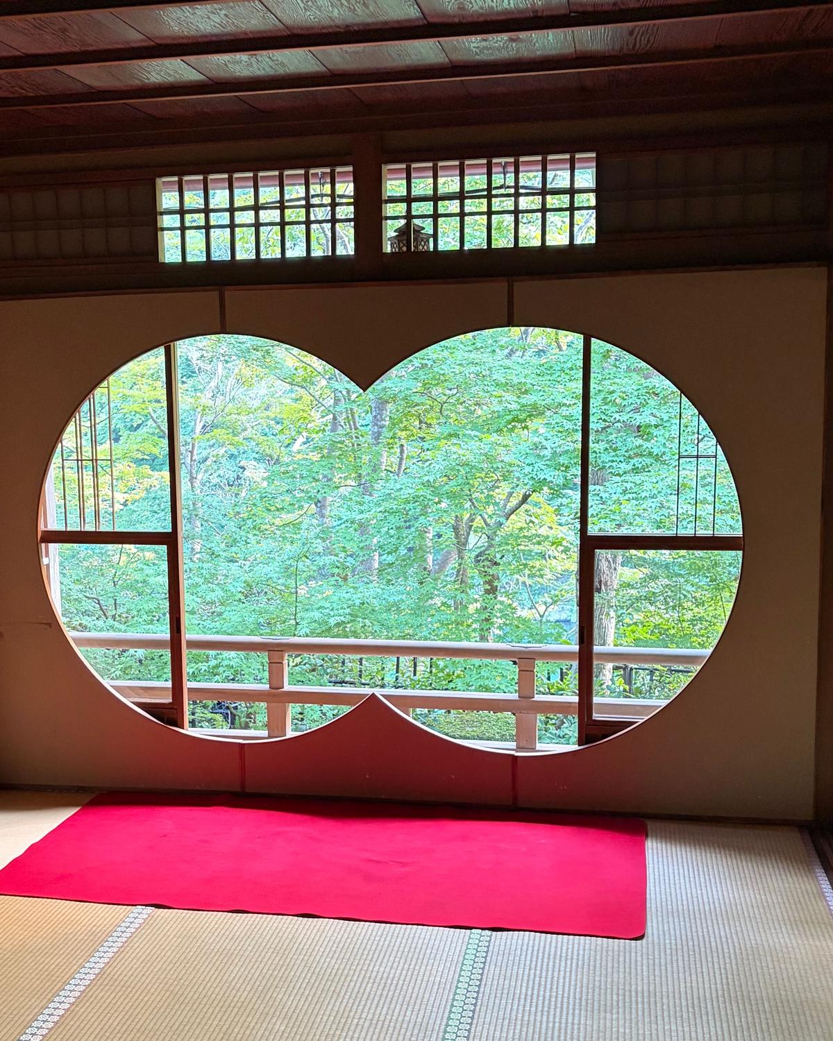 伝統的な日本の室内、木製の格子窓と丸窓から緑豊かな庭園が見える。畳の上に赤い敷物が置かれている。