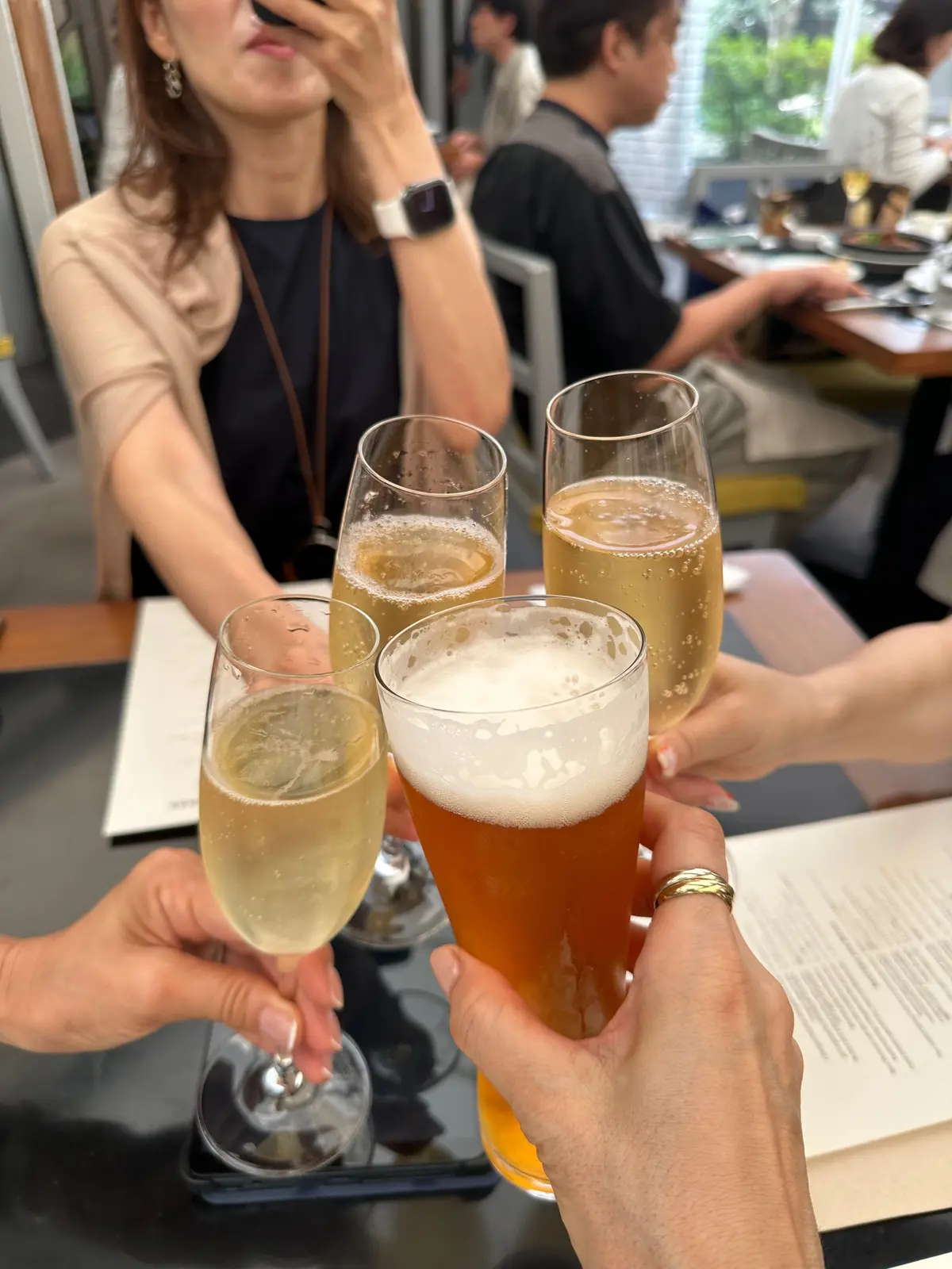 テーブルで飲み物を片手に、グラスを掲げて乾杯する人々の様子。様々な種類の飲み物が、グラスやフルートグラスに入っている。背景には、レストランのテーブル席が見える。
