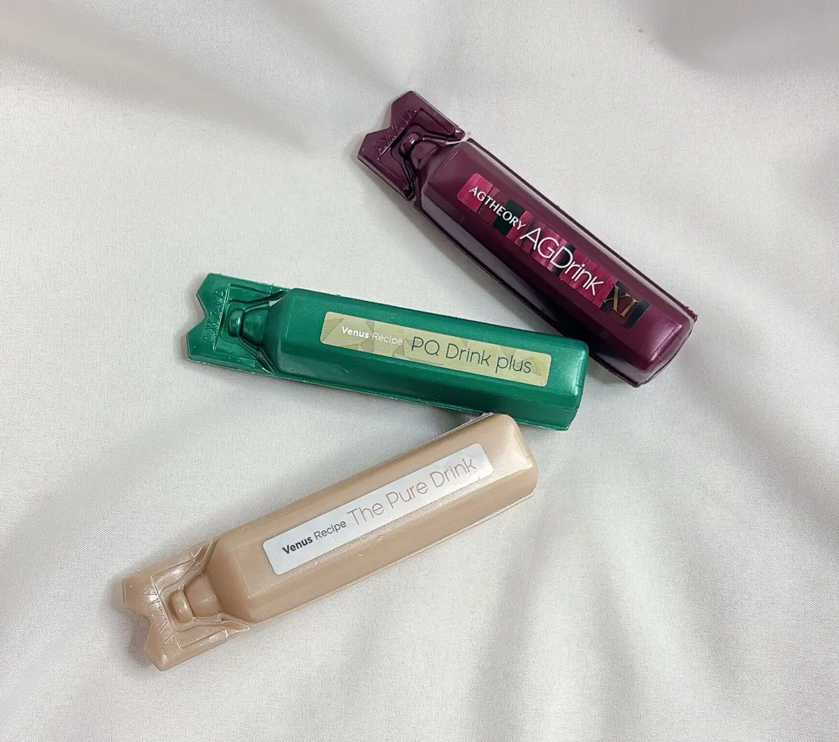 異なる色の3つのスティック状ドリンクパックが置かれている写真。上から、紫色の「AGDrink XI」、緑色の「Venus Recipe PQ Drink plus」、ベージュ色の「Venus Recipe The Pure Drink」。