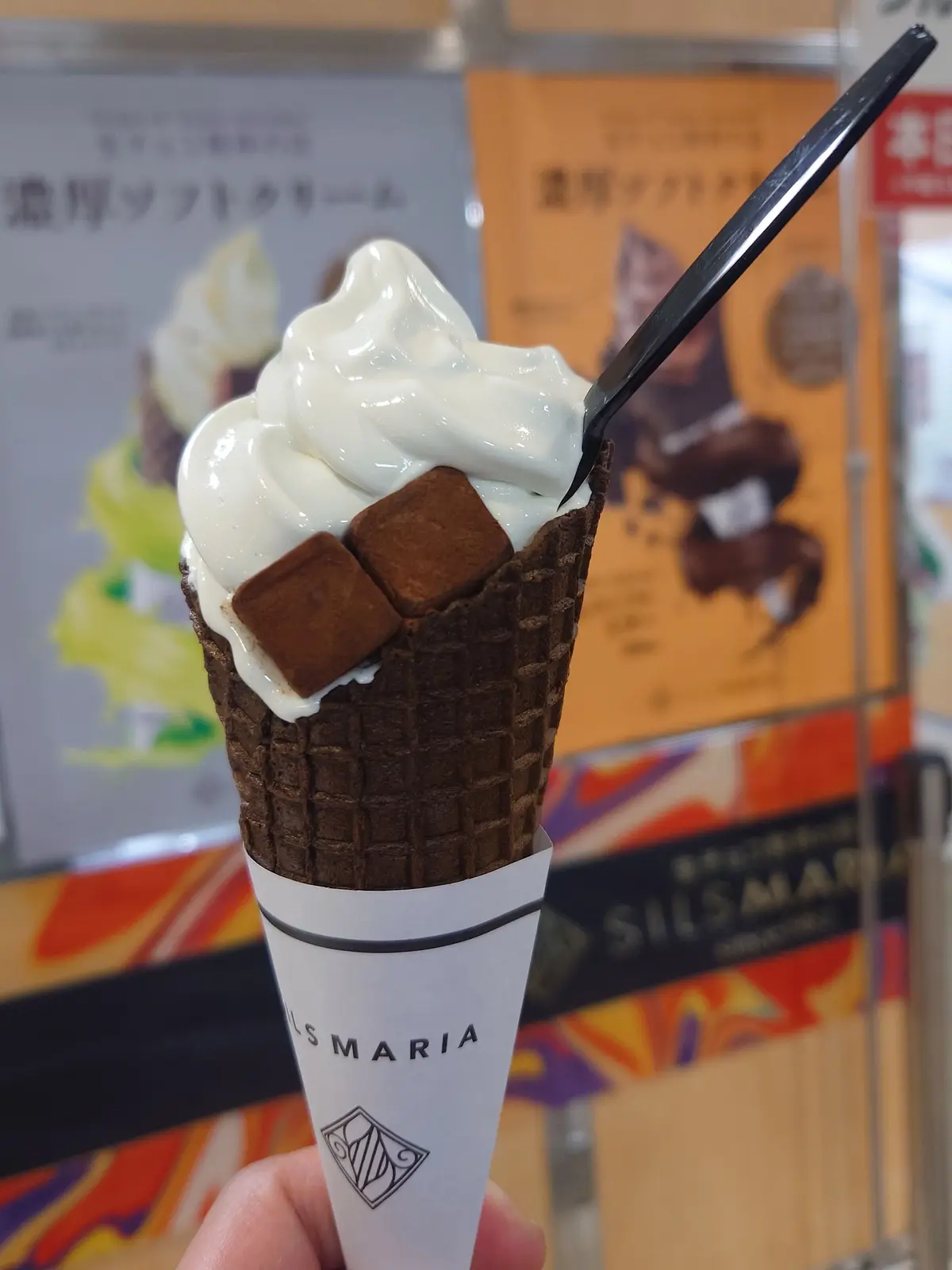 SILS MARIAのソフトクリーム。ワッフルコーンに白くてふわふわのソフトクリームが盛られ、チョコレートの角切りが散らされている。木の棒が付いており、背景にはSILS MARIAの広告が見える。