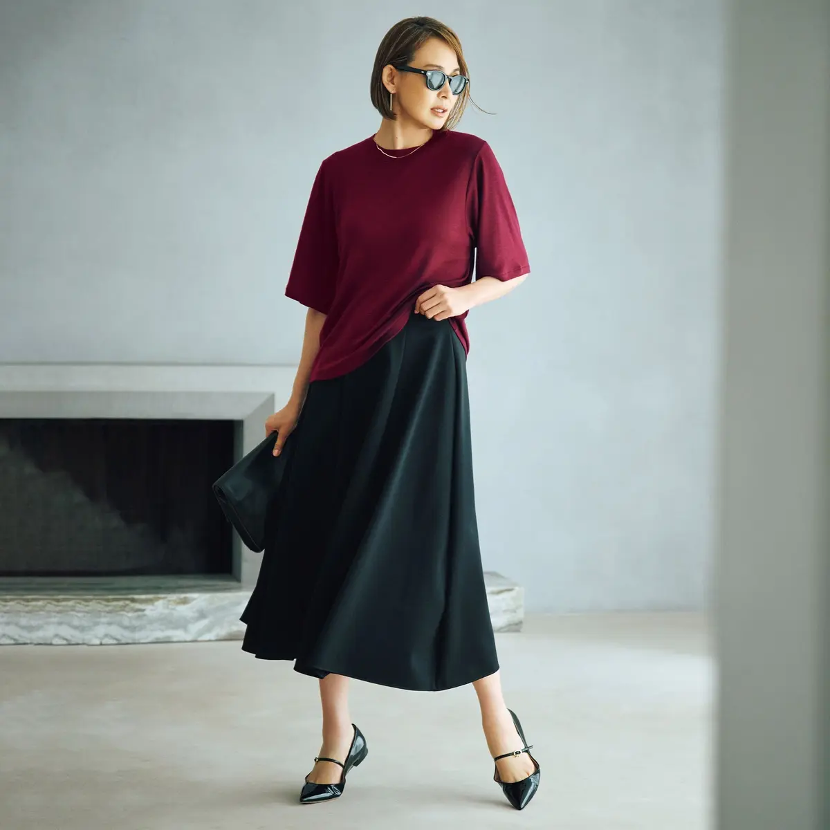 赤いTシャツと黒のミモレ丈スカートを着用した女性。黒いストラップパンプスを履き、黒縁のサングラスをかけている。背景には、コンクリートの壁と暖炉がある。