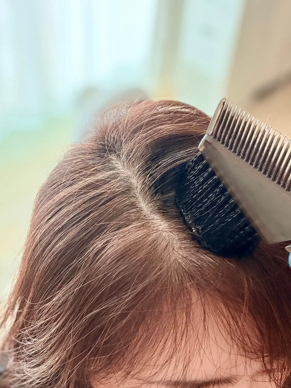 茶色の髪に、黒いヘアマスカラが塗られている様子。分け目は中央で、ヘアマスカラは根元から髪の毛に沿って塗布されている。櫛が添えられている。