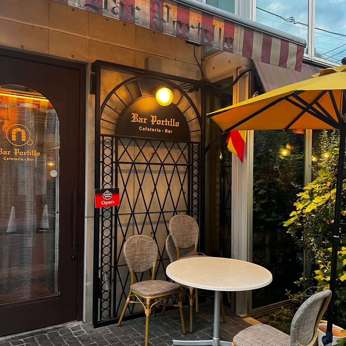 Bar Portilloの入り口。アーチ型の鉄格子ゲートが開き、カフェテリアとバーの看板が掲げられている。テーブルと椅子が並び、赤と白のストライプのオーニングが設置されている。