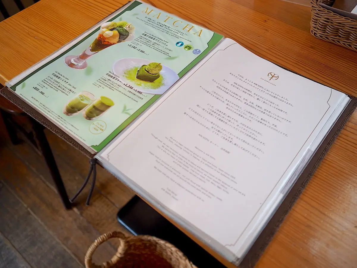 木製のテーブルに置かれた「MATCHA」と書かれたメニューブック。左ページには抹茶スイーツの写真と価格が記載され、右ページには日本語の文章が書かれている。ブックは開いており、一部がテーブルの端に掛かっている。