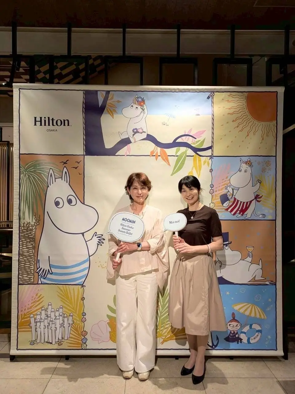 ヒルトン大阪の「ムーミンサマースイーツビュッフェ」のイベントで、2人の女性が「MOOMIN」と「Moi moi!」と書かれたボードを手に笑顔で立つ。背景にはムーミンのイラストが描かれたカラフルなバナーがある。