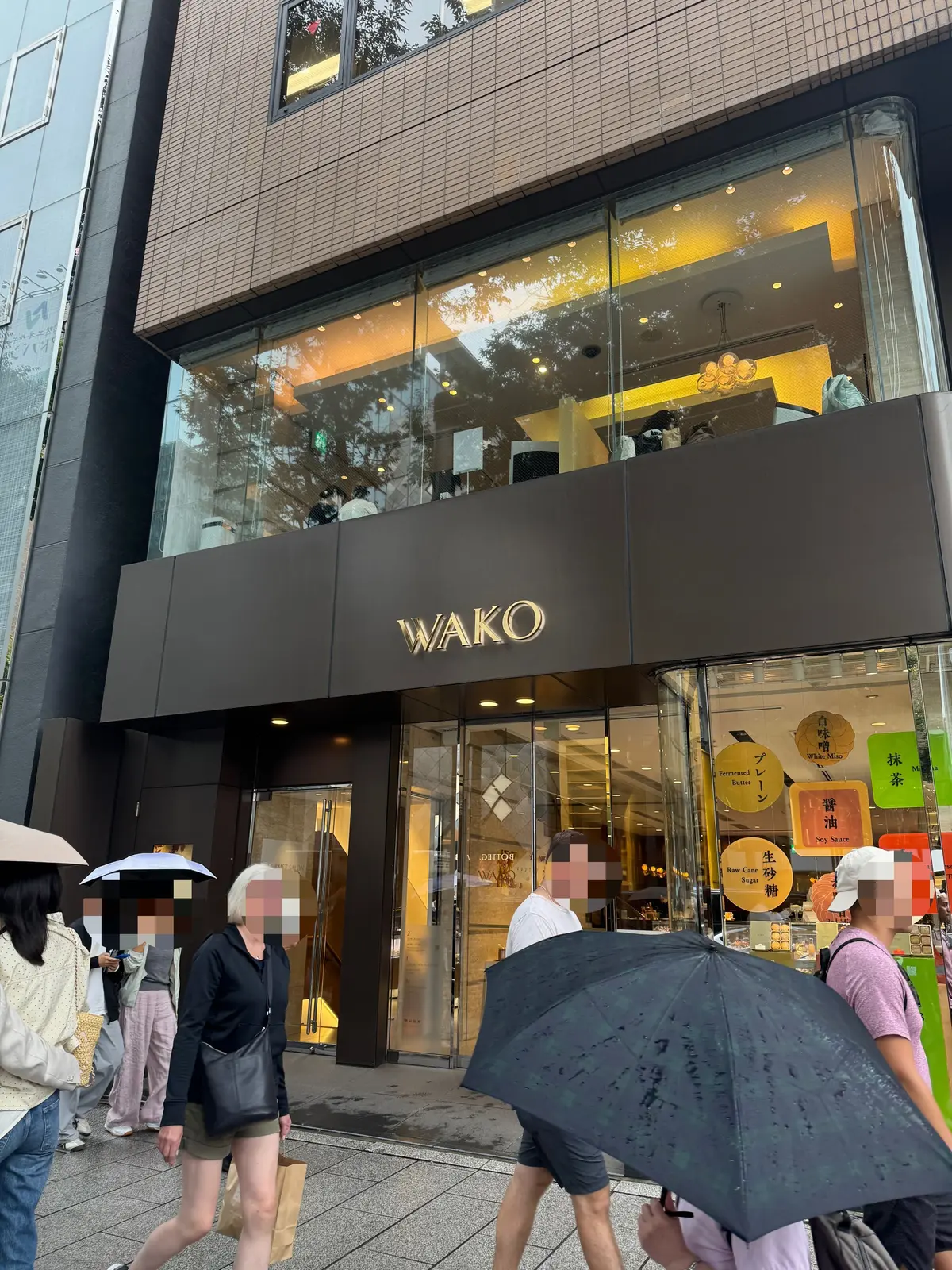 銀座のデパート「和光」の外観。ガラス張りのショーウィンドウに商品が並び、入り口付近では雨具をさした人々が通り過ぎる様子。